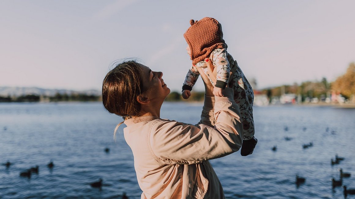 Assicurazione sulla vita: come funziona? Madre tiene sollevato il suo bimbo davanti a un lago