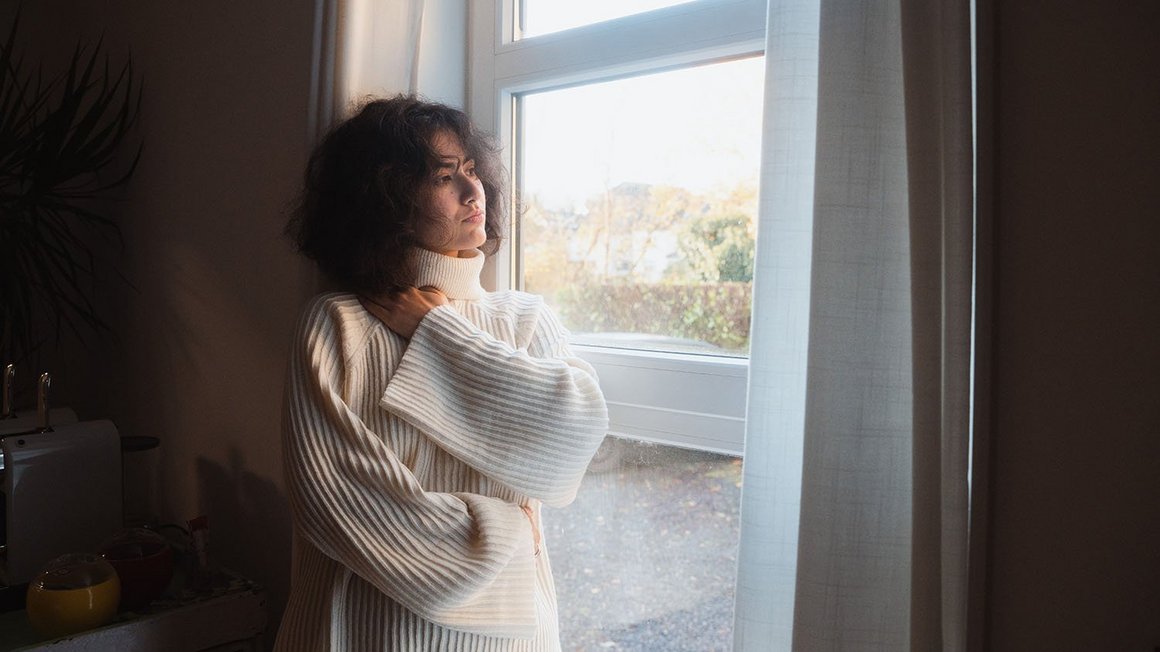 Donna con maglione bianco in piedi davanti a una finestra mentre guarda fuori pensierosa.