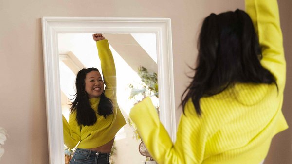 Ragazza col maglione giallo allunga euforica il braccio davanti allo specchio.